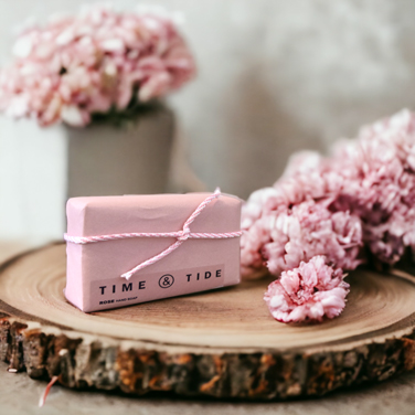 A pink soap bar tied with pink & white ribbon, on a wooden plinth with pink flowers