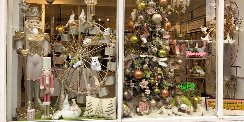 A shop window with a large pink nutcracker statue, a large standing ferris wheel decorated with festive accessories, a large snowy Christmas tree to the right on the image with large pink, light green and cream baubles, pink and green standing monkeys around base of tree and sparkling dragonfly clip ons attached to the tree.